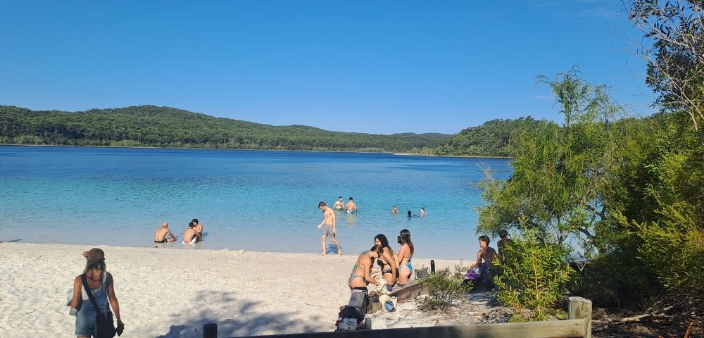 A beach scene with people swimming and relaxing near clear blue water and green hills in the background.