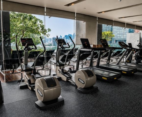 A modern gym with cardio equipment, including treadmills and ellipticals, overlooking a city skyline through large windows.