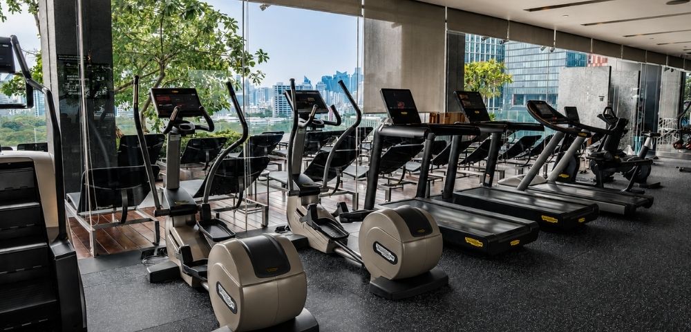 A modern gym with cardio equipment, including treadmills and ellipticals, overlooking a city skyline through large windows.