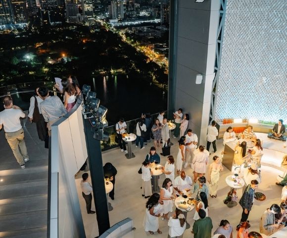 A rooftop bar scene at night with people mingling, tables with drinks, and a city skyline visible in the background.