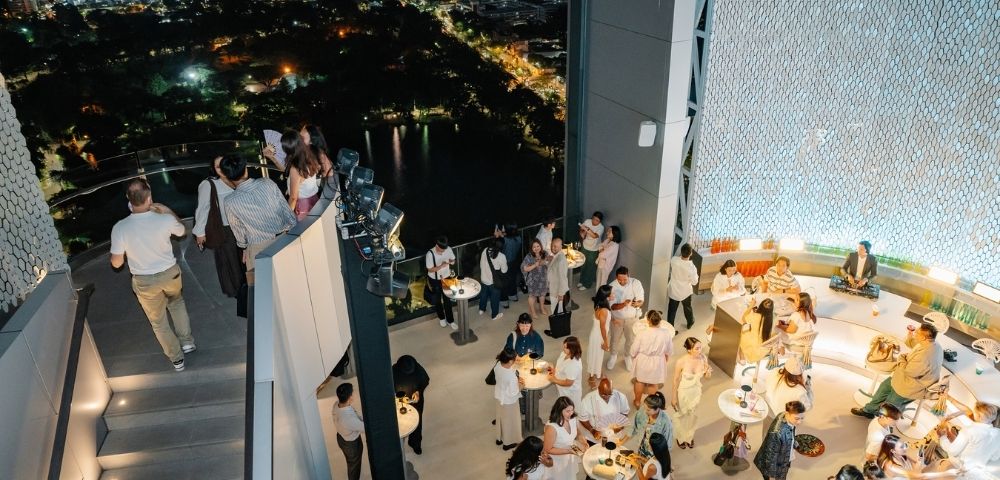 A rooftop bar scene at night with people mingling, tables with drinks, and a city skyline visible in the background.