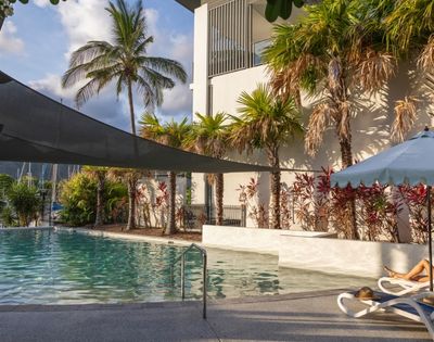 A tranquil swimming pool area surrounded by palm trees and lush greenery, with lounge chairs and a building in the background.