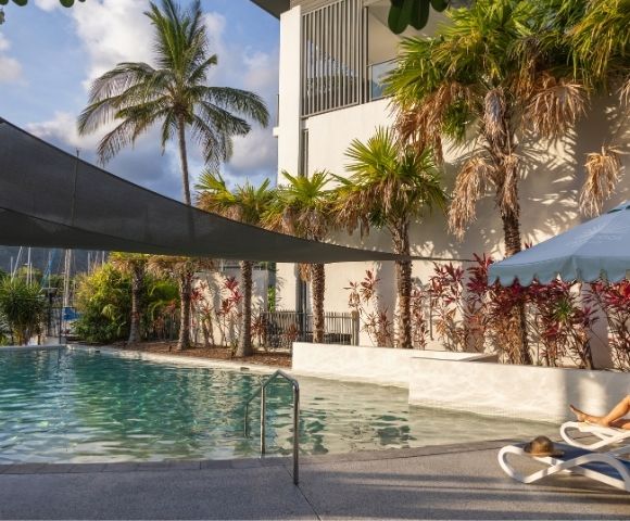 A tranquil swimming pool area surrounded by palm trees and lush greenery, with lounge chairs and a building in the background.