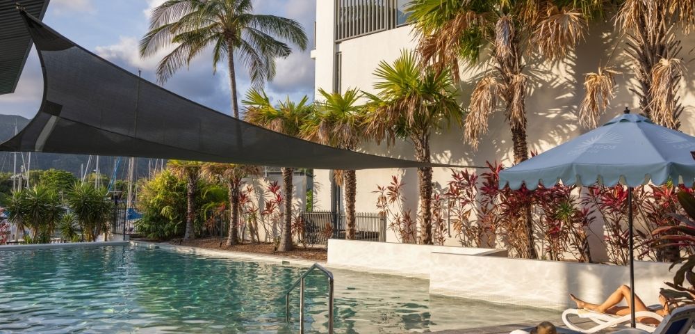 A tranquil swimming pool area surrounded by palm trees and lush greenery, with lounge chairs and a building in the background.