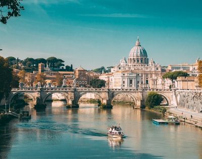A boat travels on the Tiber River with St. Peter’s Basilica and a stone bridge in the background under a clear blue sky in Rome, Italy.