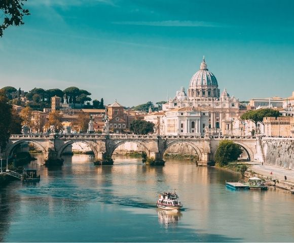 A boat travels on the Tiber River with St. Peter’s Basilica and a stone bridge in the background under a clear blue sky in Rome, Italy.