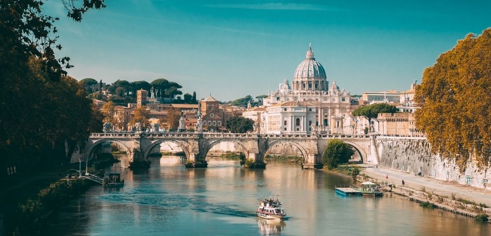 A boat travels on the Tiber River with St. Peter’s Basilica and a stone bridge in the background under a clear blue sky in Rome, Italy.