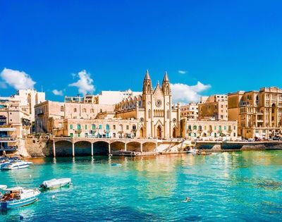 A historic church stands on the waterfront, surrounded by tan buildings, with small boats floating on clear turquoise water under a bright blue sky.
