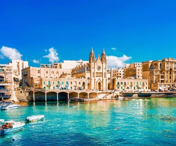 A historic church stands on the waterfront, surrounded by tan buildings, with small boats floating on clear turquoise water under a bright blue sky.