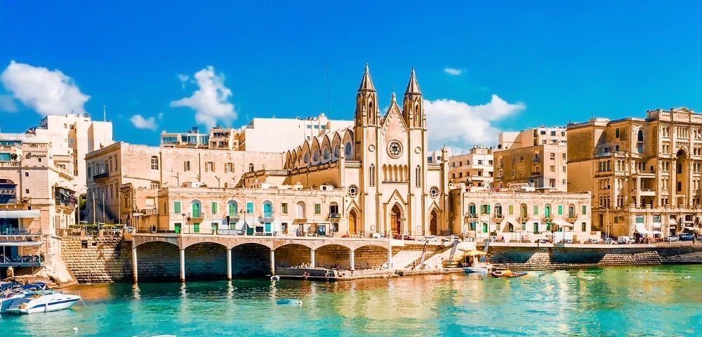 A historic church stands on the waterfront, surrounded by tan buildings, with small boats floating on clear turquoise water under a bright blue sky.