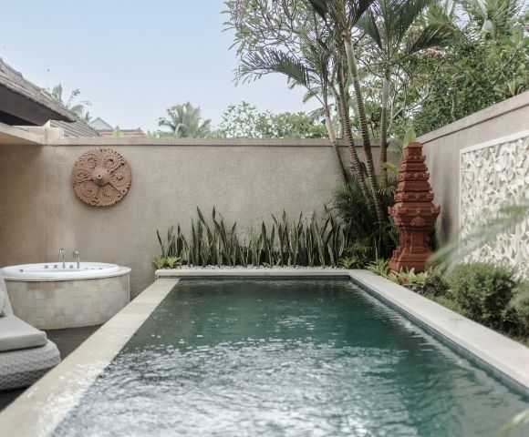 A serene backyard with a small swimming pool, a hot tub, lush greenery, and decorative stone walls.