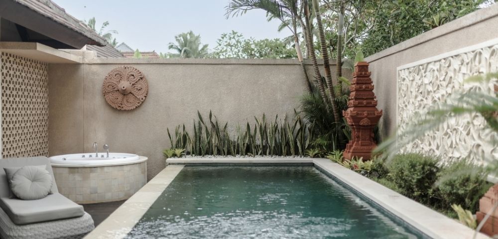 A serene backyard with a small swimming pool, a hot tub, lush greenery, and decorative stone walls.