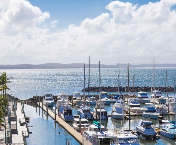 A scenic view of a marina with numerous boats and yachts docked, set against a shimmering body of water and a partly cloudy sky.