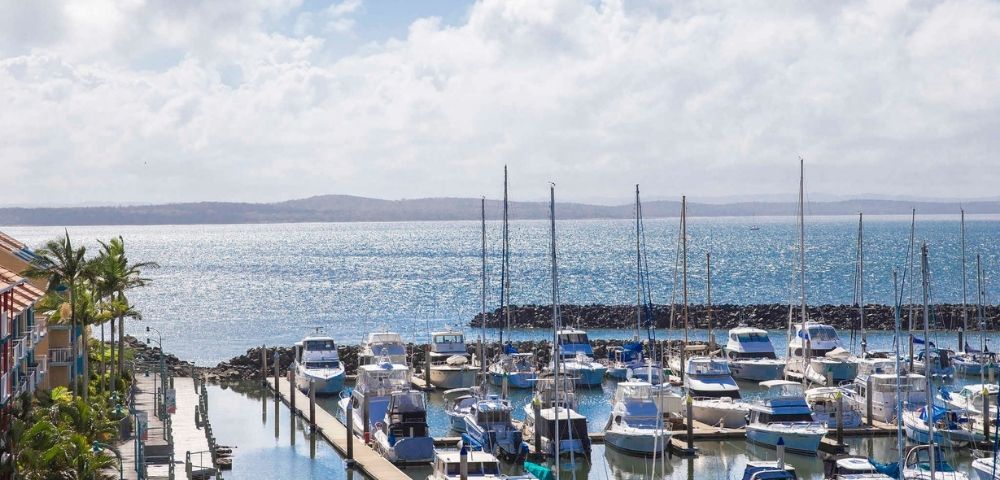 A scenic view of a marina with numerous boats and yachts docked, set against a shimmering body of water and a partly cloudy sky.
