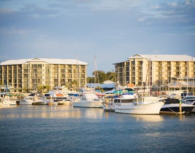 A marina scene featuring various boats docked in the water, with two multi-story buildings in the background under a cloudy sky.