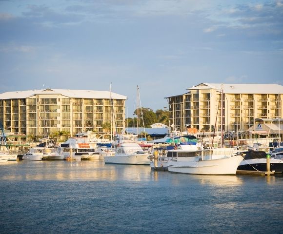 A marina scene featuring various boats docked in the water, with two multi-story buildings in the background under a cloudy sky.
