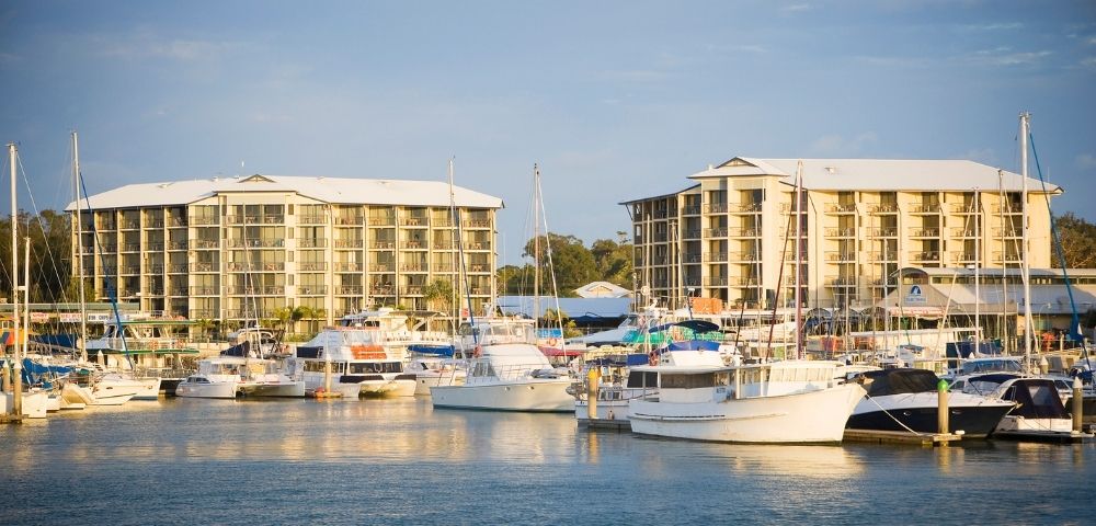 A marina scene featuring various boats docked in the water, with two multi-story buildings in the background under a cloudy sky.