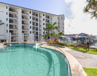 A clear swimming pool in a tropical setting, surrounded by a building with balconies and lush greenery.