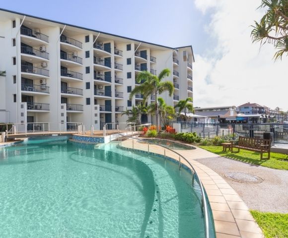 A clear swimming pool in a tropical setting, surrounded by a building with balconies and lush greenery.
