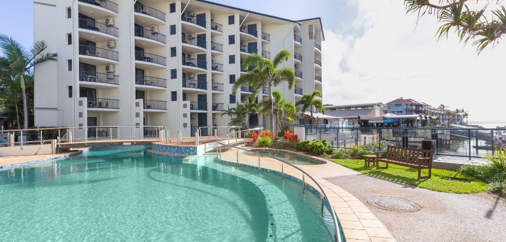 A clear swimming pool in a tropical setting, surrounded by a building with balconies and lush greenery.