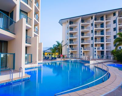 A serene hotel pool area with modern buildings surrounding it, featuring lounge chairs and palm trees under a clear sky.