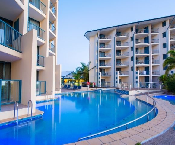 A serene hotel pool area with modern buildings surrounding it, featuring lounge chairs and palm trees under a clear sky.