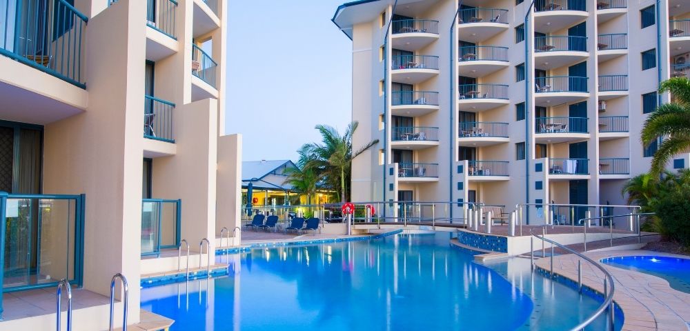 A serene hotel pool area with modern buildings surrounding it, featuring lounge chairs and palm trees under a clear sky.