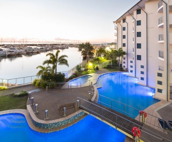 A view of a marina with boats, surrounded by a hotel building and illuminated swimming pools, during sunset.