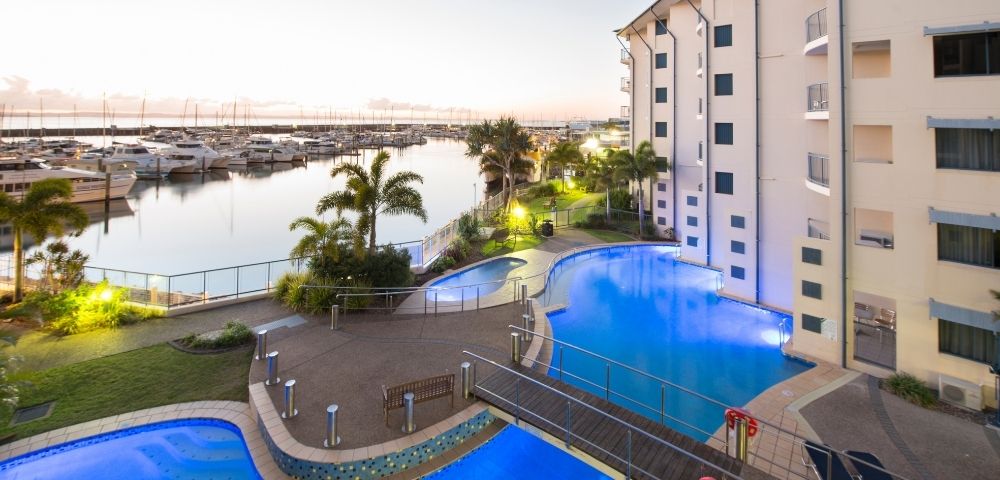 A view of a marina with boats, surrounded by a hotel building and illuminated swimming pools, during sunset.