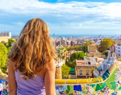 A woman with long, blonde hair faces away from the camera, looking out over the cityscape of Barcelona, Spain, from Park Güell. The foreground features a colorful mosaic bench, with unique architectural elements of the park, including a distinctive tiled tower, visible to the right. The city stretches to the horizon under a bright, partly cloudy sky.