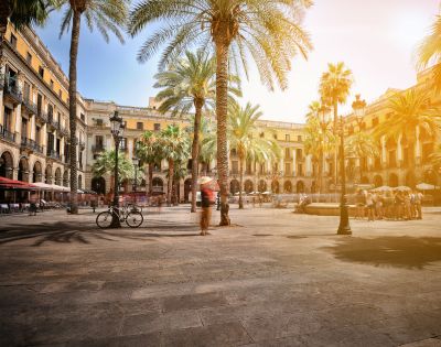 A sun-drenched European plaza with palm trees, a fountain, and blurred figures of people walking and sitting, with arcaded buildings in the background.