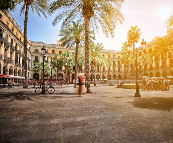 A sun-drenched European plaza with palm trees, a fountain, and blurred figures of people walking and sitting, with arcaded buildings in the background.