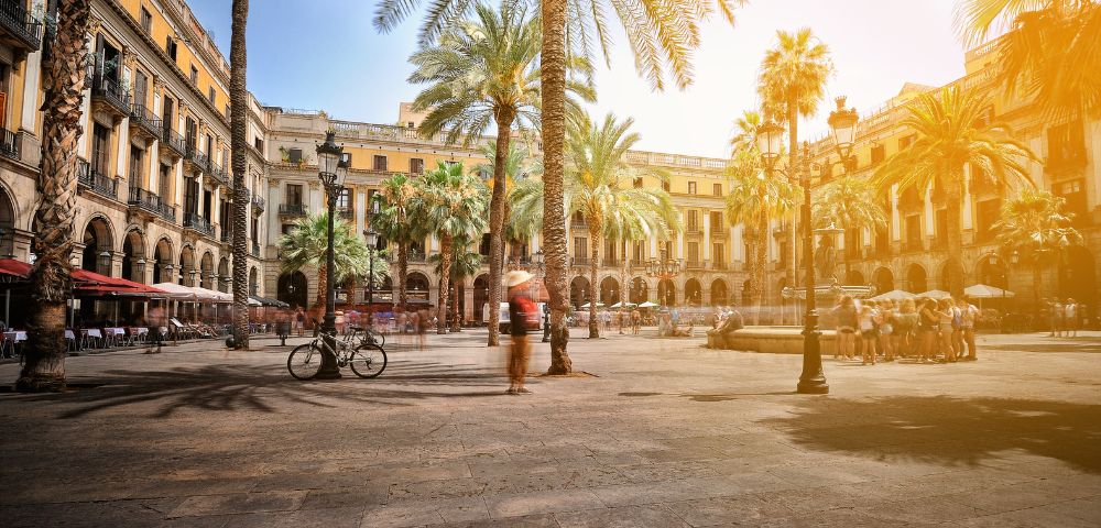 A sun-drenched European plaza with palm trees, a fountain, and blurred figures of people walking and sitting, with arcaded buildings in the background.