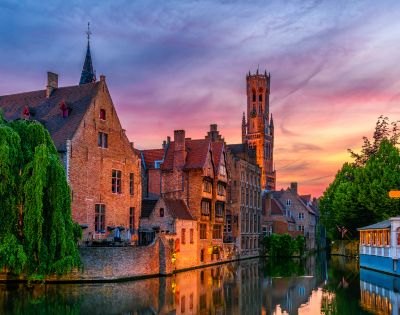 Canal in Bruges with historic brick buildings and a tall belfry tower illuminated against a vibrant sunset sky.