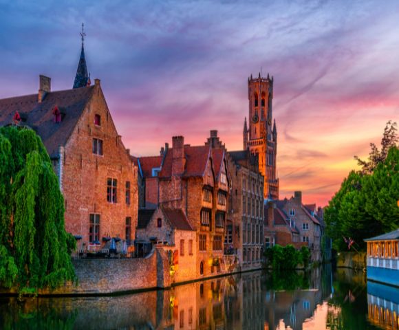 Canal in Bruges with historic brick buildings and a tall belfry tower illuminated against a vibrant sunset sky.
