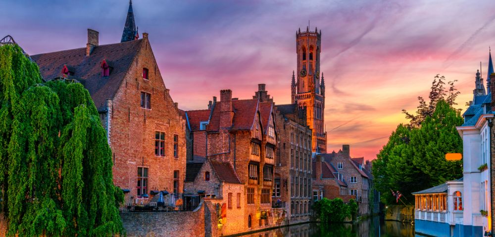Canal in Bruges with historic brick buildings and a tall belfry tower illuminated against a vibrant sunset sky.