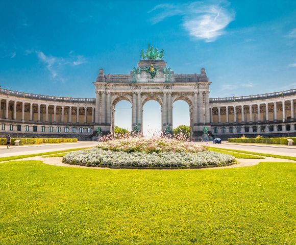 The grand Cinquantenaire Arch in Brussels with its curved colonnades, ornate sculptures, and manicured gardens under a clear blue sky.