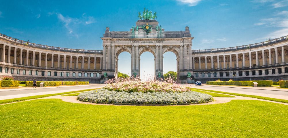 The grand Cinquantenaire Arch in Brussels with its curved colonnades, ornate sculptures, and manicured gardens under a clear blue sky.