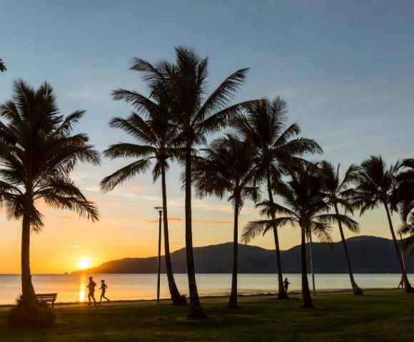 Silhouetted palm trees stand along a coastal path at sunrise, with people jogging and walking by the calm ocean and distant mountains in the back.