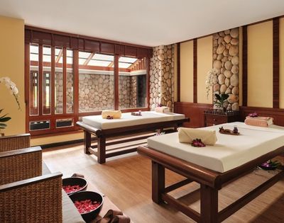 A serene spa interior featuring two massage beds with towels and flower arrangements, surrounded by wooden accents and stone walls, with natural light streaming in through large windows.