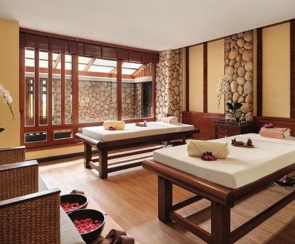 A serene spa interior featuring two massage beds with towels and flower arrangements, surrounded by wooden accents and stone walls, with natural light streaming in through large windows.