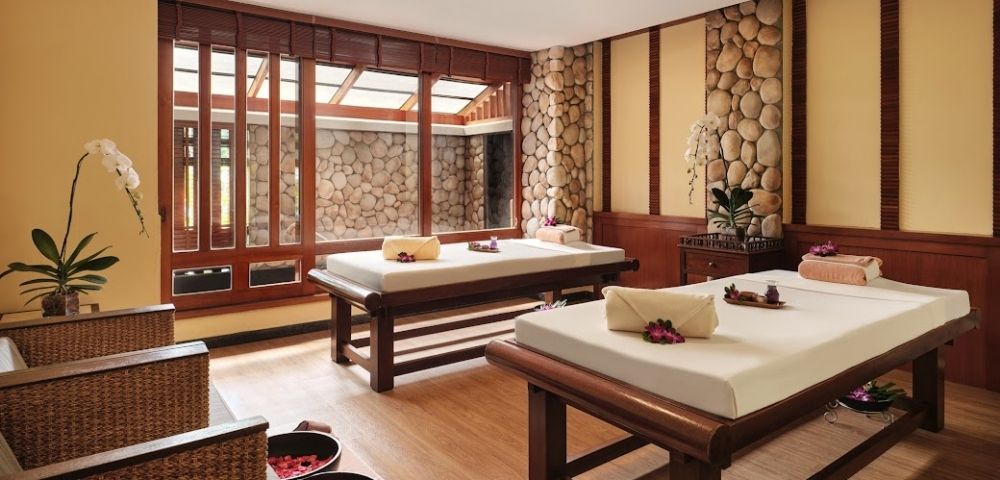 A serene spa interior featuring two massage beds with towels and flower arrangements, surrounded by wooden accents and stone walls, with natural light streaming in through large windows.