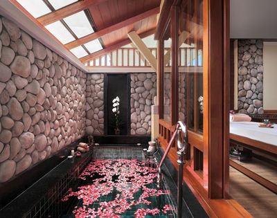 A serene spa room featuring a stone wall, a small tub filled with water and floating pink petals, and a view of an orchid plant.