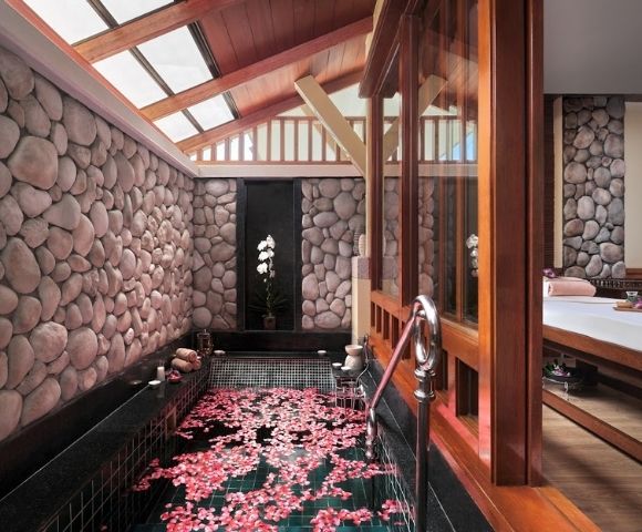 A serene spa room featuring a stone wall, a small tub filled with water and floating pink petals, and a view of an orchid plant.