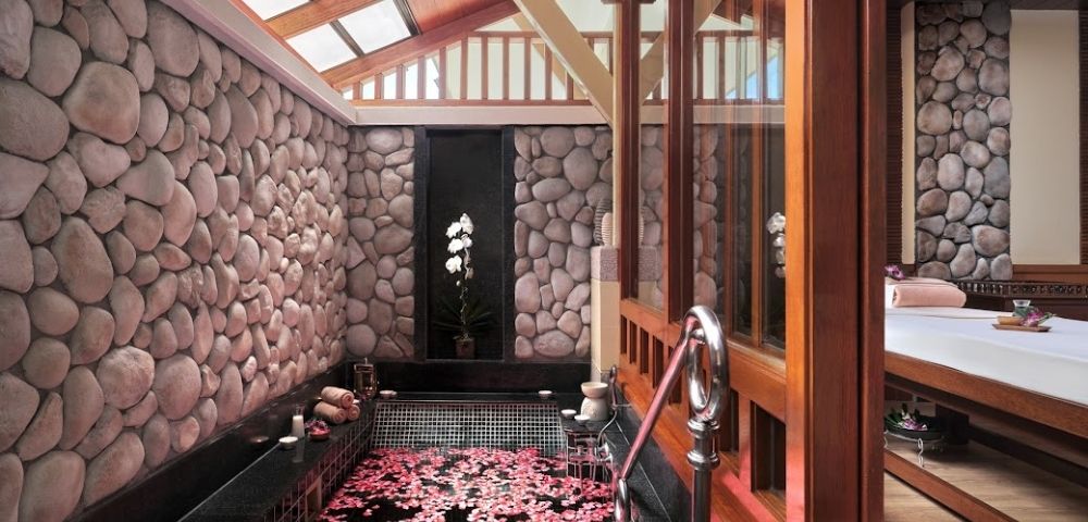 A serene spa room featuring a stone wall, a small tub filled with water and floating pink petals, and a view of an orchid plant.