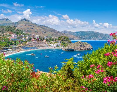 A panoramic view of a coastal town nestled on a hillside overlooking a vibrant blue bay with a small island, surrounded by lush greenery and pink flowering bushes.