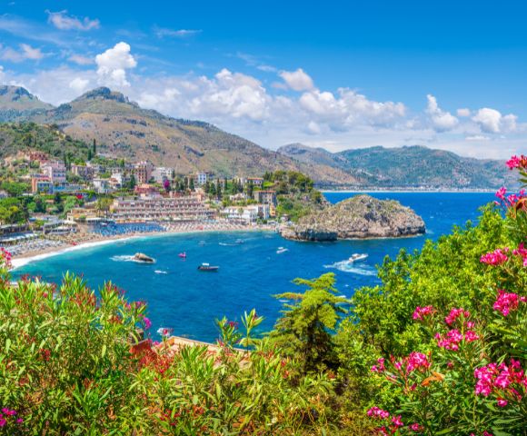 A panoramic view of a coastal town nestled on a hillside overlooking a vibrant blue bay with a small island, surrounded by lush greenery and pink flowering bushes.