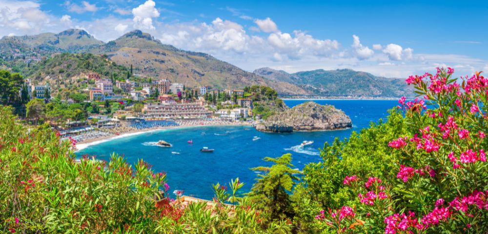 A panoramic view of a coastal town nestled on a hillside overlooking a vibrant blue bay with a small island, surrounded by lush greenery and pink flowering bushes.