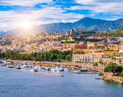 Panoramic view of a coastal city with a marina full of boats and buildings climbing a hillside under a sunny, blue sky with scattered clouds.