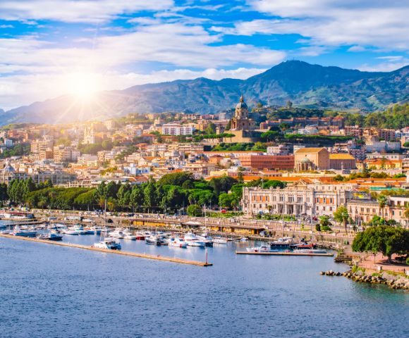Panoramic view of a coastal city with a marina full of boats and buildings climbing a hillside under a sunny, blue sky with scattered clouds.